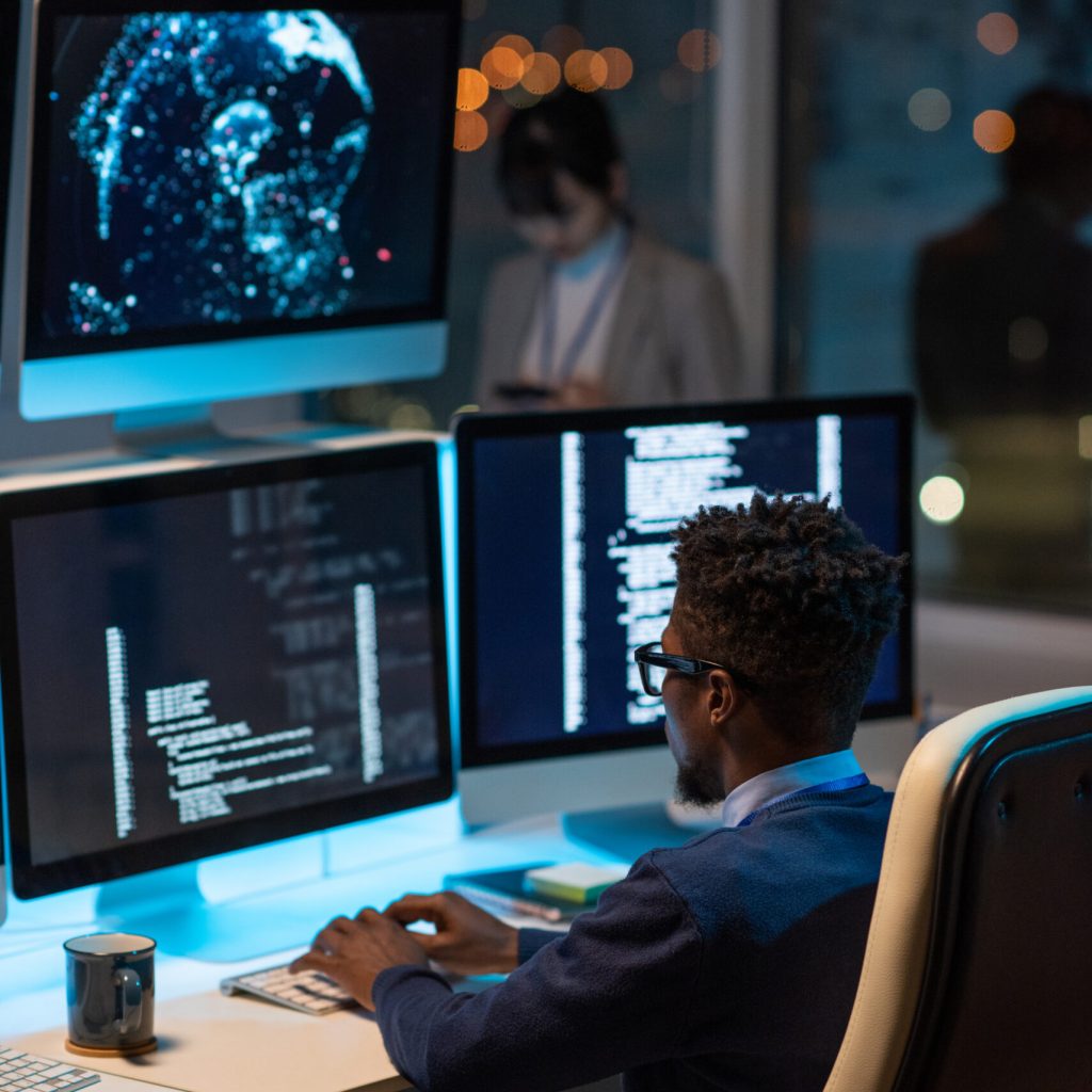 Young African developer sitting in armchair by desk and typing while looking at coded data on computer screen